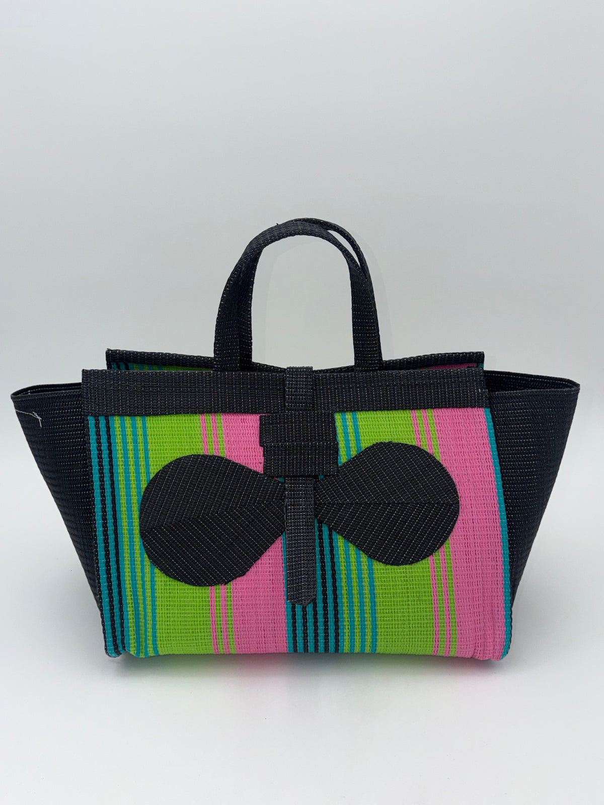 A woven tote bag with black handles and trim, featuring a front panel with vertical stripes in lime green, pink, and turquoise, accented by two large black bow shapes.